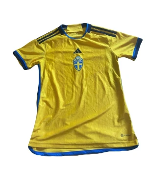 Svensk landslagströja Adidas gul - Gul svensk landslagströja från Adidas med blå detaljer och tre svarta ränder på axlarna. Broderad svensk landslagssköld på bröstet och Adidas-logga ovanför. Tillverkad i lätt och ventilerande funktionsmaterial, perfekt för fotboll eller träning.