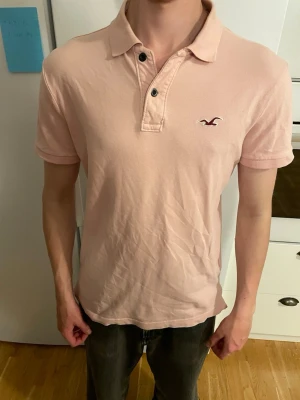 Ljusrosa piké från Hollister - Säljer en ljusrosa pikétröja från Hollister med klassisk krage och två knappar framtill. Tröjan har korta ärmar och den ikoniska Hollister-loggan broderad på bröstet. Perfekt för en avslappnad och stilren look.