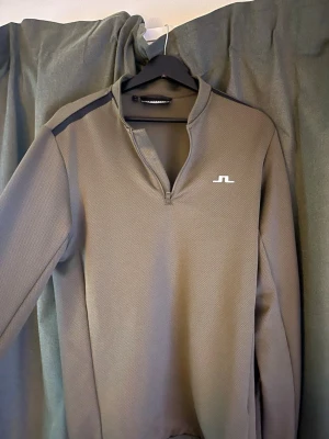 Olivgrön golftröja från J.Lindeberg - Sportig olivgrön långärmad träningströja från J.Lindeberg med halv dragkedja och diskret logga på bröstet. Tillverkad i ett tekniskt, lätt material som andas. Svart detalj på axeln ger en snygg kontrast. Perfekt för träning eller golf.
