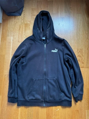Svart zip-hoodie från Puma - Svart hoodie med dragkedja från Puma. Klassisk design med huva, två stora fickor framtill och vit Puma-logga på bröstet. Mjuk bomullsblandning och ribbade muddar vid ärmslut och nederkant. Perfekt för en avslappnad streetstyle.