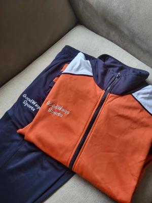 Orange och svart träningskostym GoodWays storlek L bättre XL - Sportig träningskostym från GoodWays Sports i orange och svart med vita detaljer. Jackan har dragkedja och hög krage, byxorna har resår i midjan. Perfekt för dig som gillar en färgstark och bekväm look.