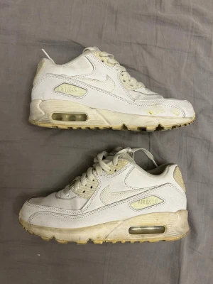 Nike Air Max 90 vita sneakers - Klassiska Nike Air Max 90 sneakers i vitt läder med beige detaljer och synlig Air-enhet i sulan. Skorna har en chunky siluett, vita skosnören och diskreta loggor på sidan och hälen. Perfekta för dig som gillar retrostil och streetwear.