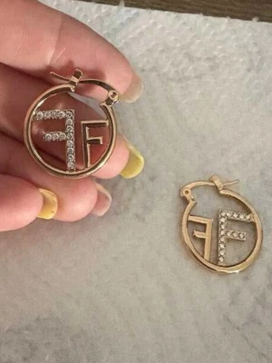 Fendi örhängen  - Säljer ett par runda guldörhängen från Fendi med FF-logga i mitten. Loggan är dekorerad med gnistrande stenar som ger extra bling. Klassisk hoop-modell med enkel låsning. Perfekta för dig som gillar exklusiva accessoarer✨