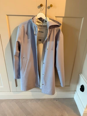 Ljusblå regnjacka från Stutterheim - Snygg ljusblå regnjacka från Stutterheim med huva och dragsko, raka linjer och tryckknappar framtill. Jackan har två snedställda fickor och är tillverkad i ett vattentätt material som håller dig torr under regniga dagar. Nypris 3000kr.