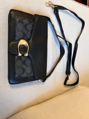 Svart axelväska från Coach - Snygg svart axelväska från Coach med justerbar rem och guldfärgat spänne. Väskan har Coachs klassiska monogrammönster på framsidan och är gjord i en mix av läder och canvas. Perfekt storlek för det viktigaste och stilren design.