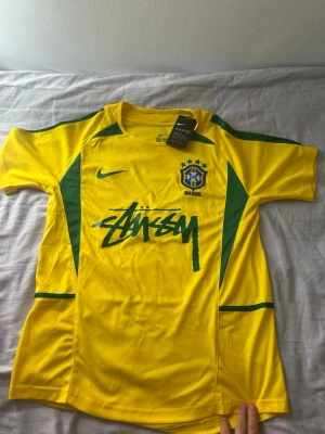 Gul Brasilien fotbollströja Nike S - Gul Brasilien fotbollströja från Nike i samarbete med Stüssy. Tröjan har gröna detaljer, korta ärmar och Brasilien-emblem på bröstet. Tillverkad i lätt och ventilerande material, perfekt för träning eller match. Stüssy-logga tryckt framtill.