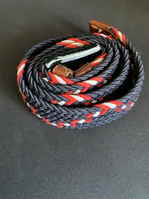 Randigt flätat bälte i marinblått - Snyggt flätat bälte med randigt mönster i marinblått, rött och vitt. Bältet har detaljer i brunt skinn och ett silvrigt spänne. Perfekt accessoar för att lyfta din outfit med lite färg och stil.