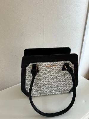 Michael Kors svartvit handväska - Snygg handväska från Michael Kors (ej äkta) i svart och vitt med klassiskt MK-mönster över hela väskan. Väskan har svarta handtag och detaljer i guld, samt en fyrkantig form som ger en modern look. Perfekt för dig som gillar stilrena accessoarer. Väskan är flitigt använd och fickorna är lite slitna. Finns flera fack som rymmer mycket grejer. Saknar de tillhörande axelremmarna. 