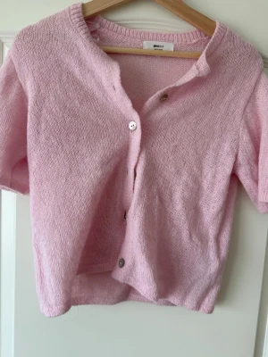 Ljusrosa stickad kofta Gina Tricot - Söt ljusrosa stickad kofta från Gina Tricot i ullblandning. Kort modell med korta ärmar och knäppning framtill med tre knappar. Perfekt att slänga över en topp för en chill look.