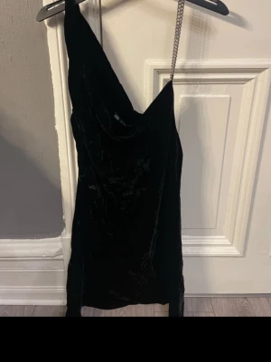 Svart sammetsklänning oneshoulder - Snygg svart kort sammetsklänning med oneshoulder-design och en kedja som axelband. Klänningen har en tight passform och är perfekt för dig som vill sticka ut. Materialet är mjukt och glansigt, vilket ger en lyxig känsla.