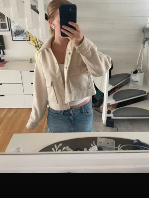 Beige cropped teddyjacka med fickor - Säljer en beige teddyjacka i croppad modell med stora bröstfickor och knappar framtill. lite smink på insidan men inget som syns när den används.