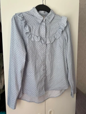 Ljusblå skjorta - Supersöt ljusblå skjorta från H&M i storlek 38 med små mörkblå prickar och volangdetalj över bröstet. Klassisk krage och knäppning framtill. Två  extra knappar medföljer!❤️