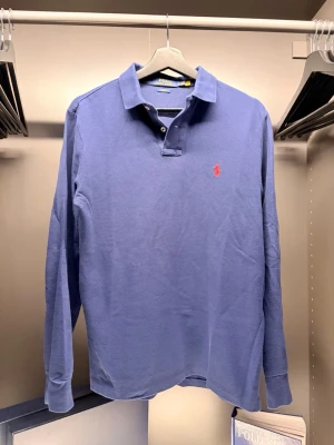  Ralph Lauren - Snygg blå långärmad skjorta från Ralph Lauren i slim fit-modell. Skjortan har klassisk krage, knappar framtill och den ikoniska röda loggan broderad på bröstet. Perfekt för dig som gillar stilrena plagg.
