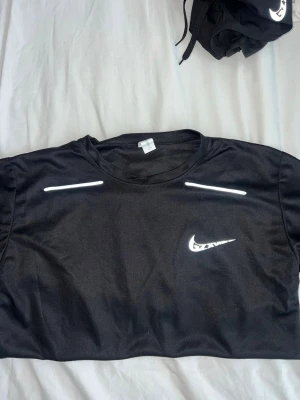 Svart Nike träningsset - Säljer ett svart träningsset från Nike med t-shirt och shorts. Båda plaggen har vita Nike-loggor och är gjorda i ett lätt och funktionellt material som passar perfekt för träning. T-shirten har rund hals och reflexdetaljer, shortsen har elastisk midja och fickor.