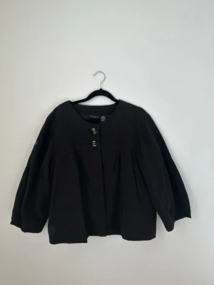 Svart jacka med trekvartsärm från Bay Studio - Stilren svart jacka från Bay Studio med rund halsringning och två knappar framtill i guld och svart. Jackan har trekvartsärmar och en lös, rak passform.