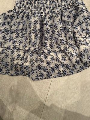 Vit och blå mönstrad kjol från H&M - Kort kjol från H&M i vitt med blått grafiskt mönster. Kjolen har resår i midjan och är tillverkad i ett lätt och luftigt material, perfekt för varmare dagar. Snygg och enkel att matcha med olika toppar.