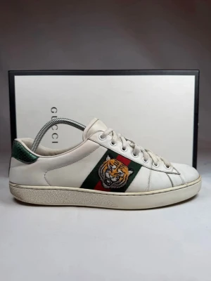 Gucci ace tiger - Gucci sneakers i vitt läder med klassiska gröna och röda ränder på sidan samt en broderad tigerpatch. Skorna har vita snören, låg profil och grön detalj på hälen. Perfekta för dig som vill sticka ut med en ikonisk och lyxig sneaker.