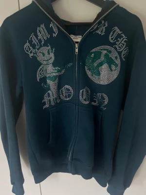 Blå hoodie Aim For The Moon - Mörkblå hoodie med dragkedja från Aim For The Moon. Framsidan har glittrande stenar i motiv av en tecknad figur och texten 'Aim For The Moon'. Utrustad med huva och kängurufickor. Perfekt för dig som gillar streetwear och unika detaljer.