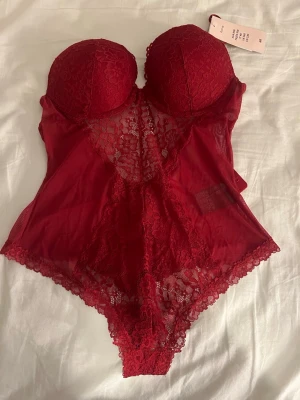 Röd spetsbody från H&M - Snygg röd body från H&M med vadderade kupor och detaljer i spets och mesh. Djup urringning och dekorativa spetskanter. Axelbanden är justerbara och bodyn har hakknäppning i grenen. Perfekt för dig som vill ha något extra till underkläderna.