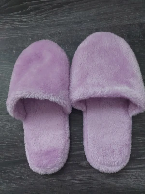 Lila fluffiga tofflor - Mysiga lila tofflor med fluffig yta och öppen häl. Tofflorna har en mjuk ovandel i syntetiskt material och en vit sula med greppmönster. Perfekta för att hålla fötterna varma hemma. Storlek 42.