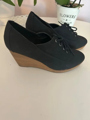 Svarta kilklackar med snörning - Snygga svarta skor med kilklack och snörning framtill. Ovandelen är i ett matt material och klacken har en trälook. Perfekta för dig som vill ha både höjd och stabilitet. Passar till många olika stilar och outfits.