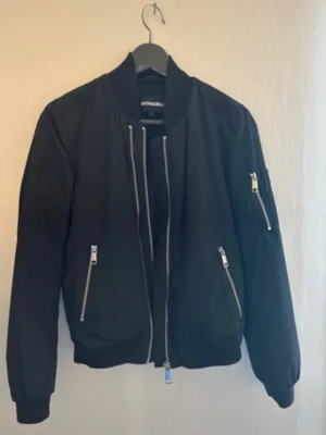 RockandBlue Svart bomberjacka med silverdetaljer - Säljer en svart bomberjacka från RockandBlue med silverfärgade dragkedjor och ribbade muddar. Jackan har en klassisk passform och en ficka med dragkedja på ärmen. Perfekt för dig som vill ha en stilren och cool look.