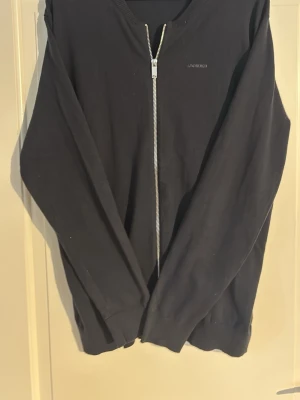 Svart ziptröja från Lindbergh - Snygg svart ziptröja från Lindbergh med relaxed fit. Tröjan har hel dragkedja framtill, diskret logga på bröstet och ribbade muddar vid hals, ärmslut och nederkant. Perfekt till lager på lager och enkel att matcha med olika outfits.