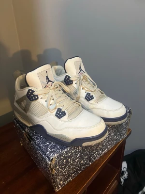 Air Jordan 4 Retro White Navy - Snygga Air Jordan 4 Retro sneakers i vitt läder med navyblå detaljer och meshpaneler. Klassisk Jumpman-logga på tungan och hälen. Skorna har en robust siluett och coola plastdetaljer vid snörningen. Perfekt för dig som gillar streetstyle och ikoniska sneakers.