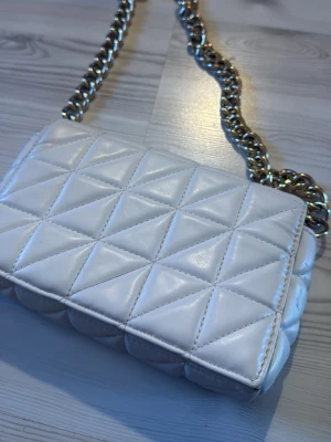 Vit quiltad handväska med kedja - Snygg vit handväska med quiltat mönster och en chunky guldfärgad kedja som axelrem. Väskan har en rektangulär form och är tillverkad i ett glansigt material som liknar konstläder. Perfekt accessoar för att lyfta din outfit.