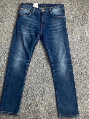 Blå jeans från Nudie med snygga fades. - Säljer ett par helt nya blå jeans från Nudie Jeans med fet tvätt. Jeansen har fem fickor, lapparna kvar, orange sömmar och en patch med logga bak i midjan. Tillverkade i mjuk denim med en cool tvättad look. Priset kan diskuteras 👍 