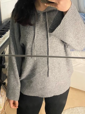 Grå stickad hoodie - Mysig grå stickad hoodie med huva och dragsko. Tröjan har breda ribbade muddar vid ärmslut och nederkant, samt en avslappnad passform. Perfekt för dig som gillar en enkel och stilren look. Materialet känns mjukt och värmande.