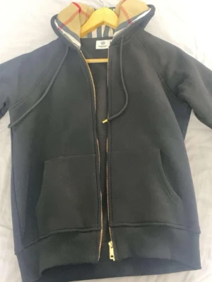 Svart hoodie med Burberry-detaljer - Snygg svart hoodie med dragkedja från Burberry. Klassisk huva med det ikoniska Burberry-rutiga mönstret på insidan och justerbara snören. Två stora fickor framtill och guldfärgade dragkedjedetaljer. Perfekt för en trendig och avslappnad stil.