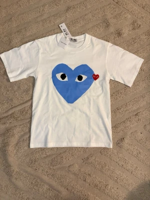 Vit Comme des Garçons Play t-shirt - Vit t-shirt från Comme des Garçons Play med stort blått hjärta med ögon tryckt på bröstet och ett litet rött broderat hjärta bredvid. Klassisk passform och rund hals. Kom dm vid minsta lilla!