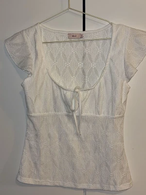 Vit spetsblus med knyt från Nelly - Söt vit blus från Nelly med spetsdetaljer och knyt framtill. Blusen har korta ärmar och en rundad urringning. Perfekt för en fräsch och somrig look. Endast testad, väldigt fint skick. Storlek M