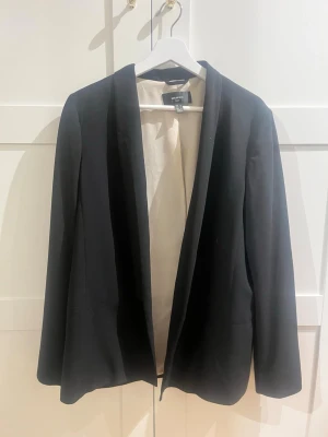 Svart kavaj från H&M med slät design - Stilren svart kavaj från Mango med öppen front och mjukt, ljust foder. Kavajen har en klassisk, rak passform och långa ärmar. Perfekt för dig som vill ha en enkel men snygg look. Materialet känns mjukt och följsamt. Passar alla storlekar beroende på hur man vill den ska sitta. 