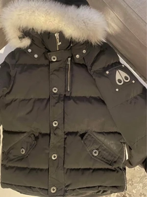 Svart dunjacka med pälskrage Moose Knuckles - Säljer en svart dunjacka från Moose Knuckles med stor fluffig pälskrage och silverfärgade detaljer. Jackan har flera fickor med knappar och dragkedjor samt logotyp på ärmen. Perfekt för kalla vinterdagar.