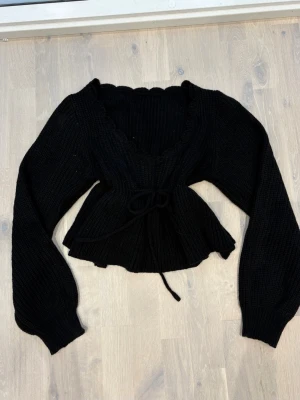 Svart stickad omlottopp - Snygg svart stickad omlottopp med knytband i midjan och volangkant nedtill. Tröjan har långa puffärmar och ribbstickat material som ger en trendig och bekväm look. Perfekt att styla med jeans eller kjol för en cool outfit.