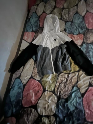 Nike vindjacka med huva, svart/vit/grå - Nike vindjacka i färgerna svart, vit och grå. Jackan har huva, dragkedja framtill och elastisk nederkant. Klassisk Nike-logga på bröstet och sportig design med färgblock. Perfekt för dig som gillar streetwear och sportig stil.