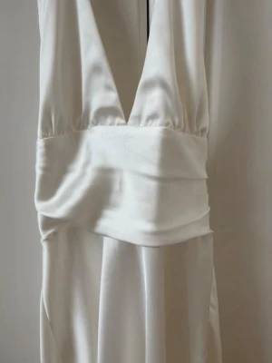 Vit halterneckklänning i satin - Säljer en vit kort halterneckklänning i satin med djup v-ringning och bred midjedetalj. Klänningen har en elegant, glansig finish och är ärmlös med axelbandslös design. Perfekt för sommarens alla tillfällen!