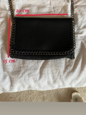 Svart axelväska med kedjerem - Snygg svart axelväska i läderimitation med silverfärgad kedjerem och kedjedetaljer runt kanterna. Aldrig använd 