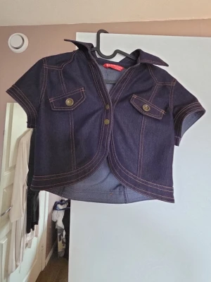 Mörkblå bolero med fickor - Snygg bolero i mörkblå denim med kontrastsömmar och två bröstfickor med knappar. Jackan har korta ärmar, markerad krage och en croppad passform som ger en trendig look. Perfekt att slänga över en t-shirt för en cool stil.