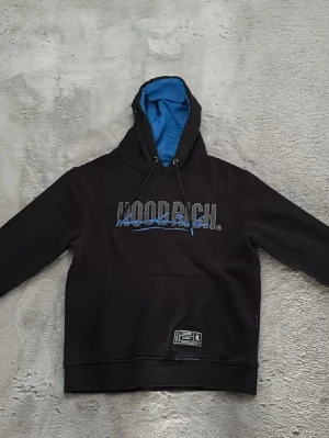 Svart HOODRICH hoodie med blå detaljer - Svart hoodie från HOODRICH med blått foder i huvan och blå logga på bröstet. Klassisk känguruficka framtill och justerbar dragsko i huvan. Perfekt för en streetwear-look och riktigt skön passform.