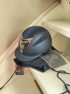 Mörkblå OneK hjälm med blå stenar - Snygg mörkblå hjälm från One K med glittrande blå stenar på ovansidan och silverdetaljer framtill. Ordinarie pris runt 3500kr. Skal strl 0 alltså så passar en liner 47-54. Aldrig använd och helt ny med lapp, har ingen liner som ingår men har en utbytbar topp-panel som ingår så man kan byta(se sista bilden). Hjälmen har en modern design och kommer med i en påse. Perfekt för dig som vill sticka ut i stallet. Skriv om du undrar något eller har några frågor!