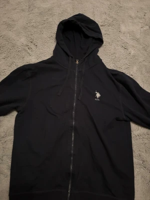 Svart zip-hoodie från USPA - Svart hoodie med dragkedja från USPA. Klassisk modell med huva och dragsko, samt broderad logga på bröstet. Tillverkad i mjukt material och har långa ärmar. Perfekt för en avslappnad stil.