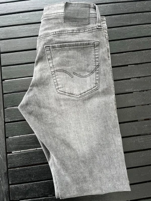 Gråa raka jeans från Jack and Jones - Säljer ett par gråa jeans från Jack and Jones  med rak passform och klassisk femficksdesign. Jeansen har en diskret broderad detalj på bakfickan och är tillverkade i mjuk denim. Perfekta för en avslappnad och stilren look. Storlek W30 L32 modell Slim Glenn 