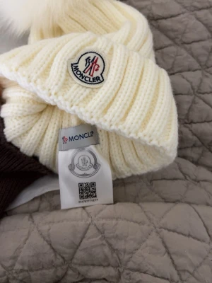 Vit ribbstickad mössa från Moncler - Snygg vit ribbstickad mössa från Moncler med uppvikt kant och stor fluffig pälstofs på toppen. Klassisk Moncler-logga framtill. Perfekt accessoar för kalla dagar och ger en lyxig touch till din vinterstil.