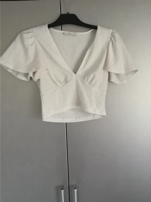 Vit croppad topp från Pigalle - Snygg vit croppad topp från Pigalle med v-ringning och korta, vida ärmar. Tillverkad i ett strukturerat tyg som ger en trendig känsla. Perfekt att matcha med höga jeans eller kjol för en fräsch look.