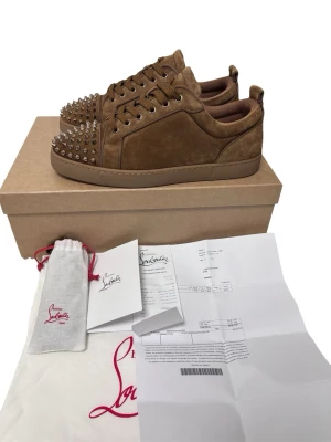 Christian Louboutin  - Säljer ett par bruna sneakers från Christian Louboutin i mocka med coola silverfärgade nitar på tån. Skorna har lågt skaft, matchande bruna skosnören och klassisk röd sula. Perfekta för dig som vill sticka ut med en lyxig och edgy stil.