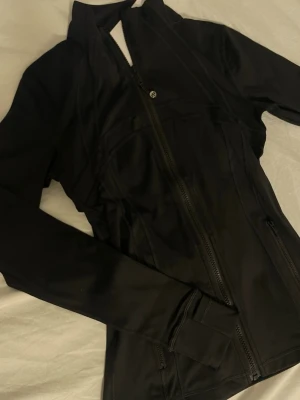 Svart sportig vindjacka med dragkedja st 4  - Svart, figurnära jacka med hel dragkedja framtill och höga muddar vid ärmsluten. Jackan har en sportig look och är tillverkad i ett slätt syntetmaterial. Perfekt för dig som gillar en clean och stilren stil.