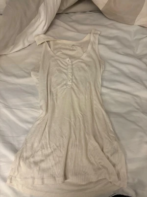 Vit ribbad linnetopp med knappar - Säljer en enkel och snygg vit ribbad linnetopp med knappdetaljer framtill. Toppen är ärmlös och har en rundad halsringning. Perfekt att matcha med jeans eller kjol för en clean look.